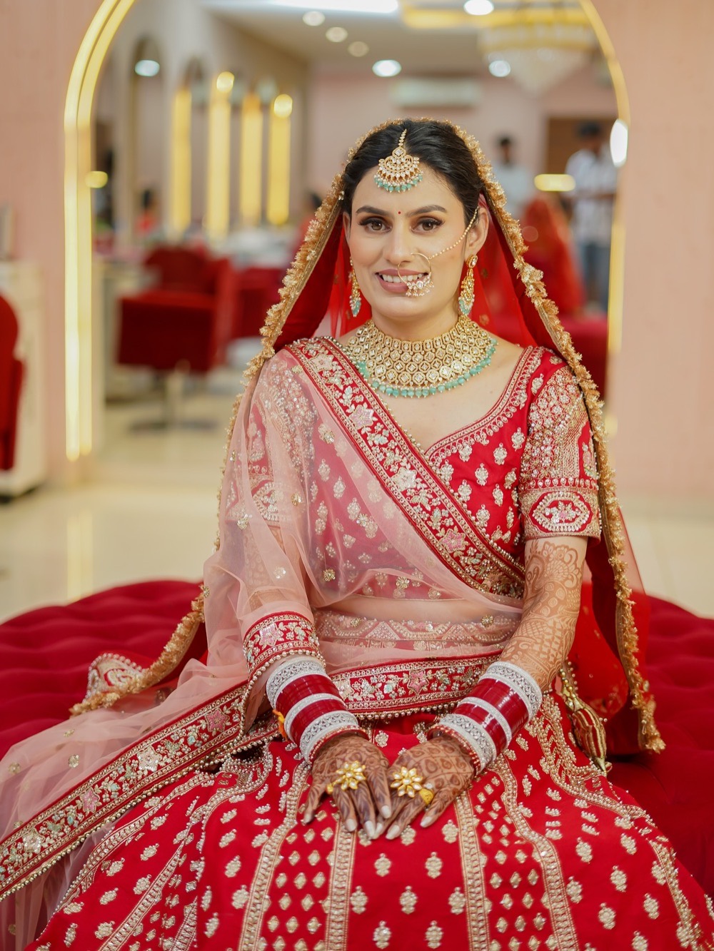 Seated bride in a red lehenga with full bridal jewellery, chooda and mehendi, makeup by Fashion World Palampur